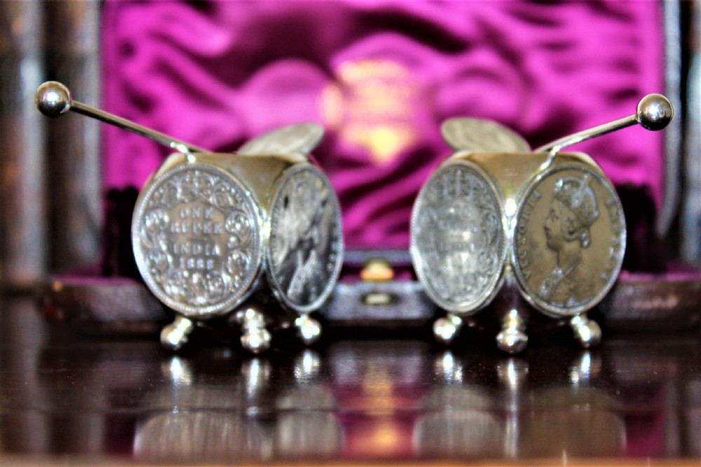 A pair of Victorian silver coin salt cellars with hinged coin lids and spoons sitting in front of an open fitted magenta velvet box. View V