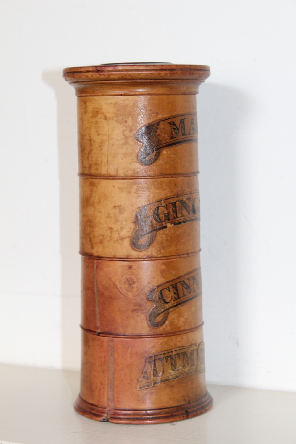 A turned wooden four-tier cylindrical spice tower with paper label banners marking Mace, Ginger, Cinnamon, and Nutmegs. View VI