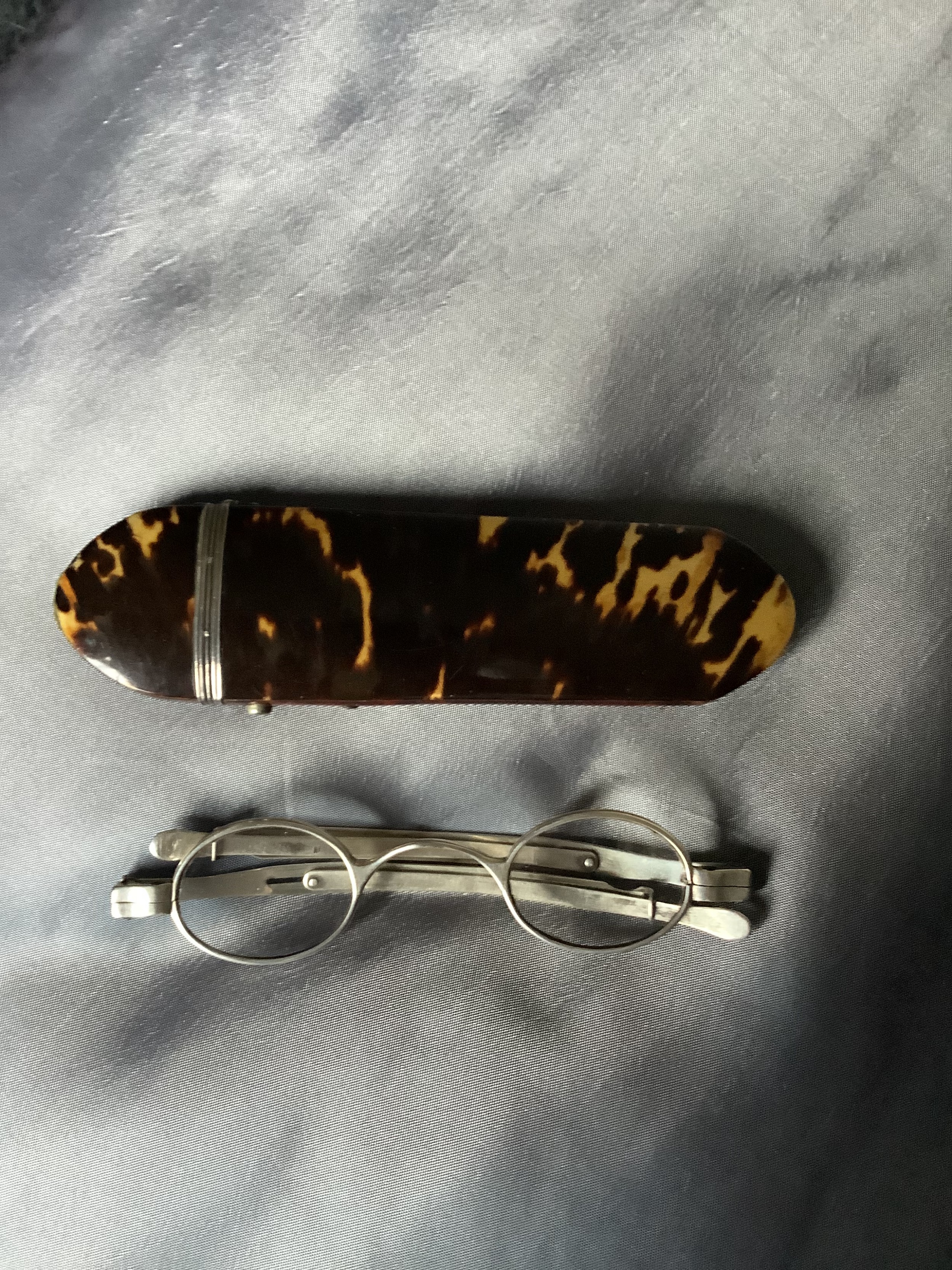 A pair of silver hallmarked folding spectacles positioned below a fitted tortoiseshell case with silver mounts.