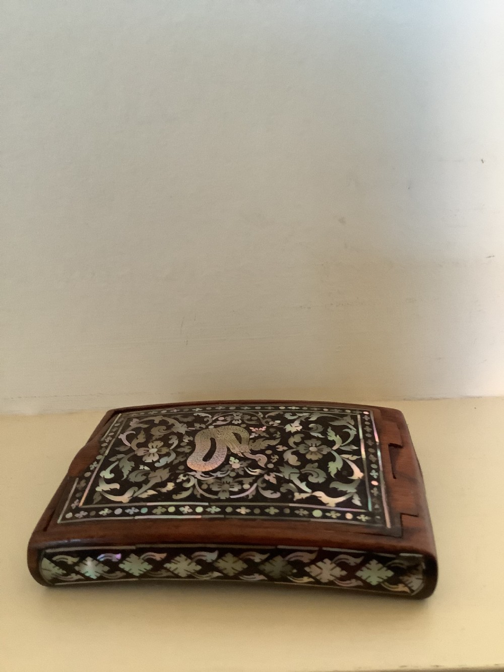 A rectangular dark hardwood box decorated with intricate mother-of-pearl inlay depicting a central serpent surrounded by floral scrolls and a geometric border. View X