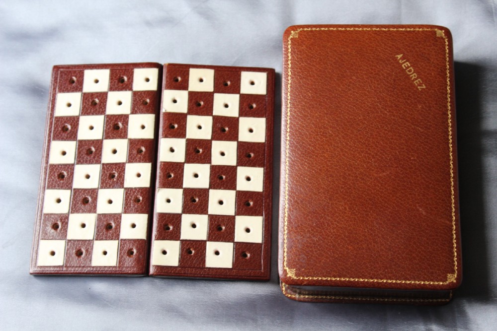 An antique miniature travelling chess set featuring a folding pegged board with alternating brown and cream squares beside a gilt-tooled brown leather case stamped Ajedrez.