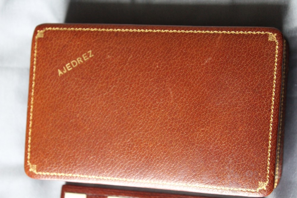 An antique miniature travelling chess set featuring a folding pegged board with alternating brown and cream squares beside a gilt-tooled brown leather case stamped Ajedrez. View II