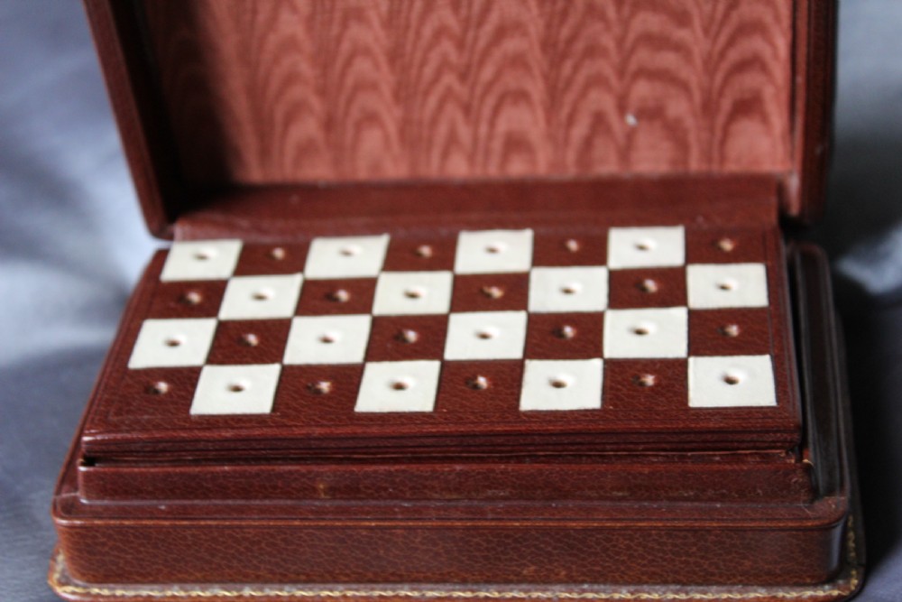An antique miniature travelling chess set featuring a folding pegged board with alternating brown and cream squares beside a gilt-tooled brown leather case stamped Ajedrez. View IV