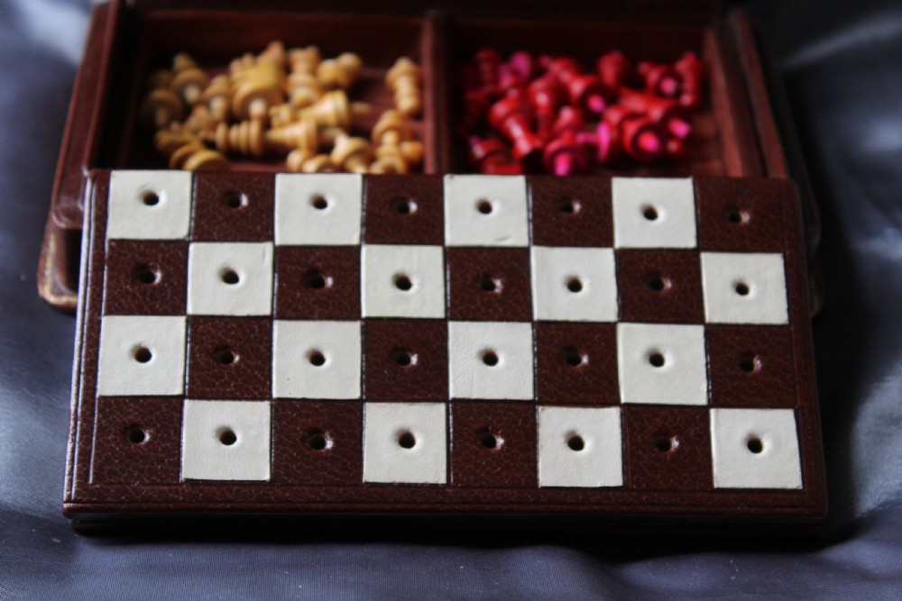 An antique miniature travelling chess set featuring a folding pegged board with alternating brown and cream squares beside a gilt-tooled brown leather case stamped Ajedrez. View VIII