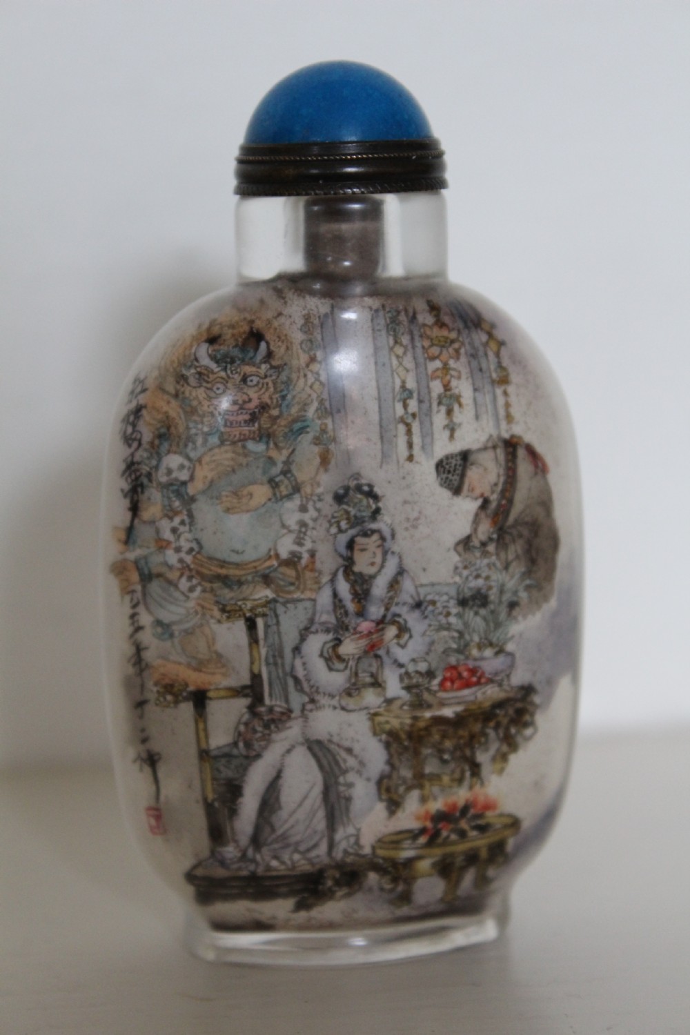 An inside-painted clear glass Chinese snuff bottle featuring detailed figures and calligraphy, topped with a rounded blue cabochon stopper. View XIV