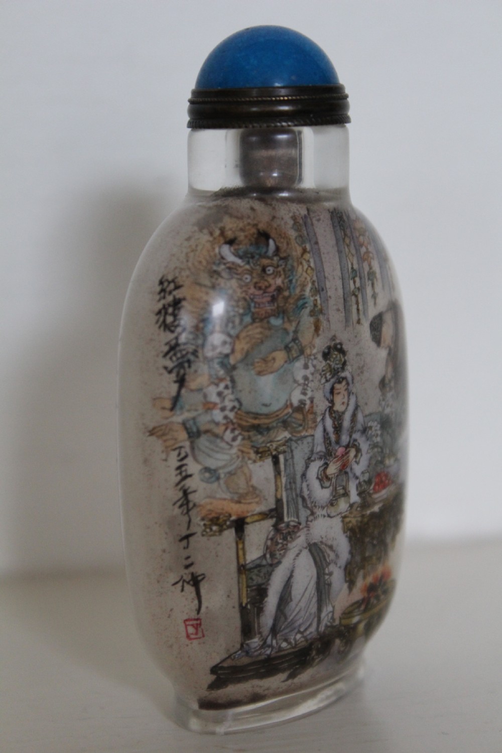 An inside-painted clear glass Chinese snuff bottle featuring detailed figures and calligraphy, topped with a rounded blue cabochon stopper. View IV