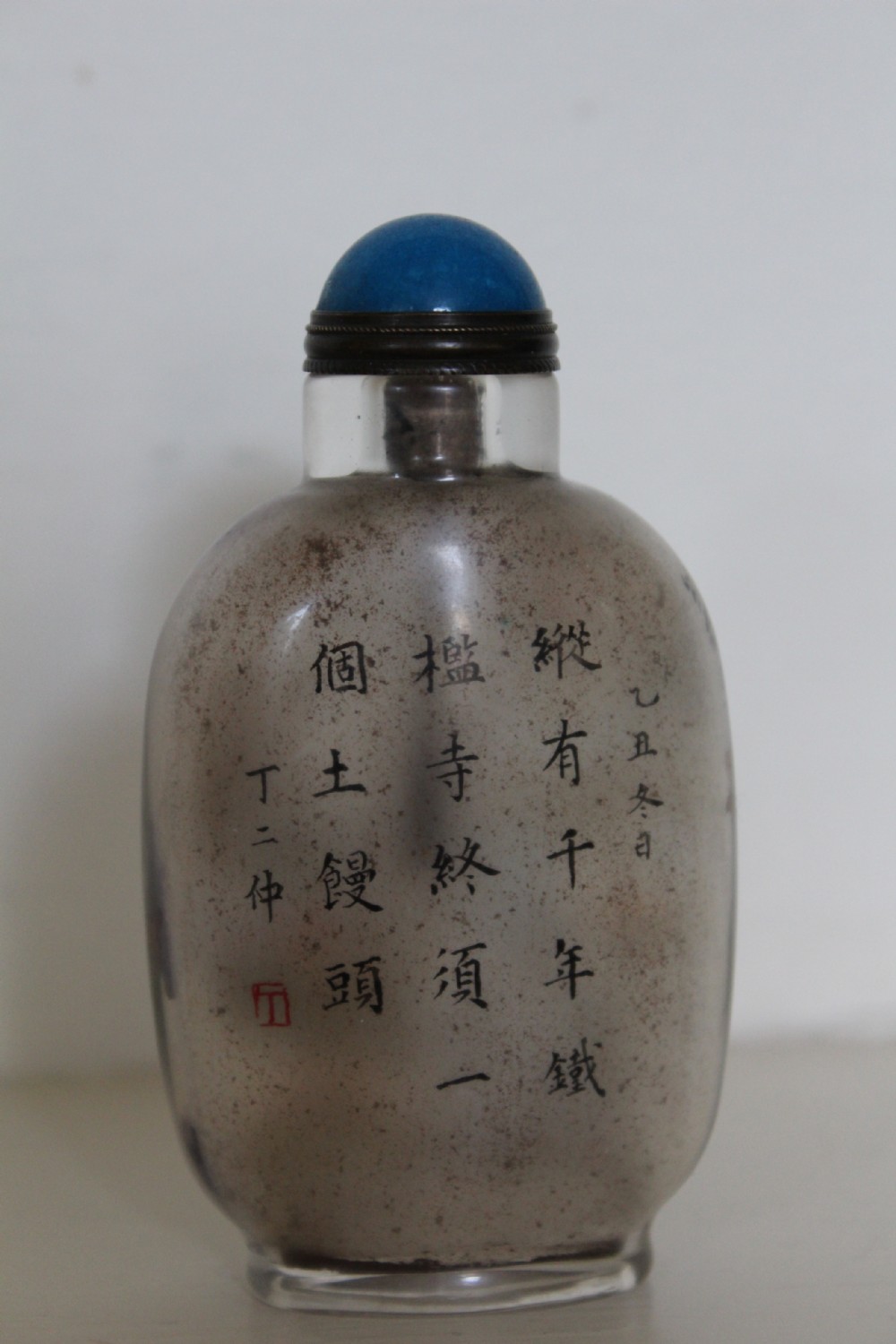 An inside-painted clear glass Chinese snuff bottle featuring detailed figures and calligraphy, topped with a rounded blue cabochon stopper. View VI