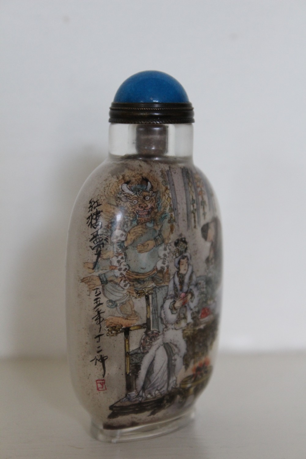An inside-painted clear glass Chinese snuff bottle featuring detailed figures and calligraphy, topped with a rounded blue cabochon stopper. View VII