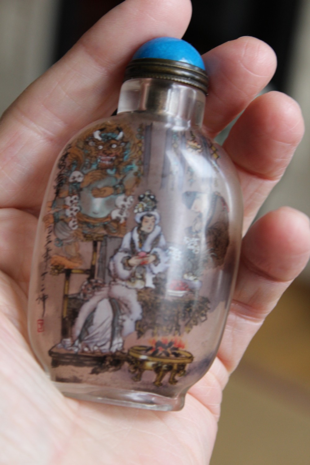 An inside-painted clear glass Chinese snuff bottle featuring detailed figures and calligraphy, topped with a rounded blue cabochon stopper. View IX