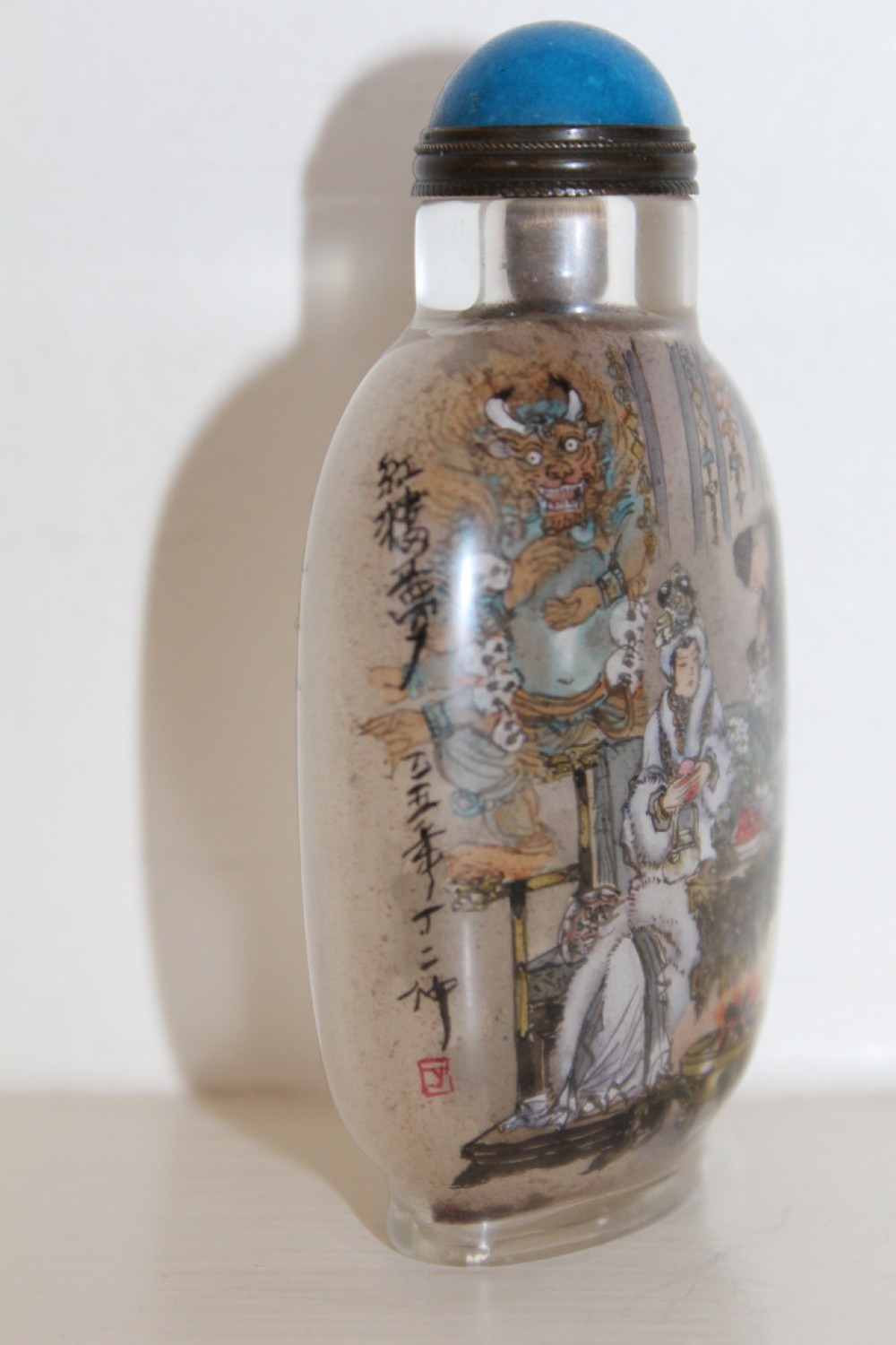 An inside-painted clear glass Chinese snuff bottle featuring detailed figures and calligraphy, topped with a rounded blue cabochon stopper. View X