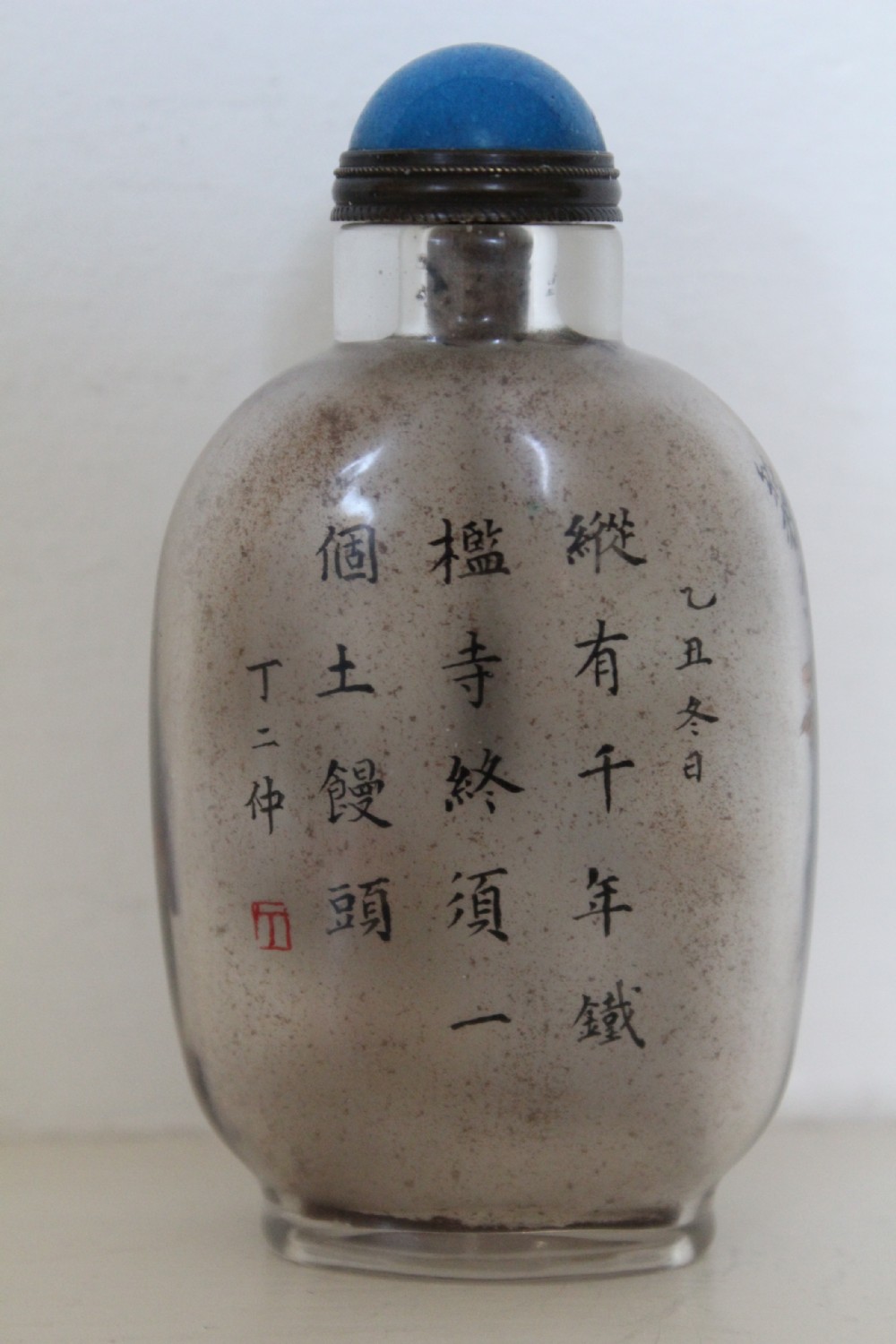 An inside-painted clear glass Chinese snuff bottle featuring detailed figures and calligraphy, topped with a rounded blue cabochon stopper. View XII
