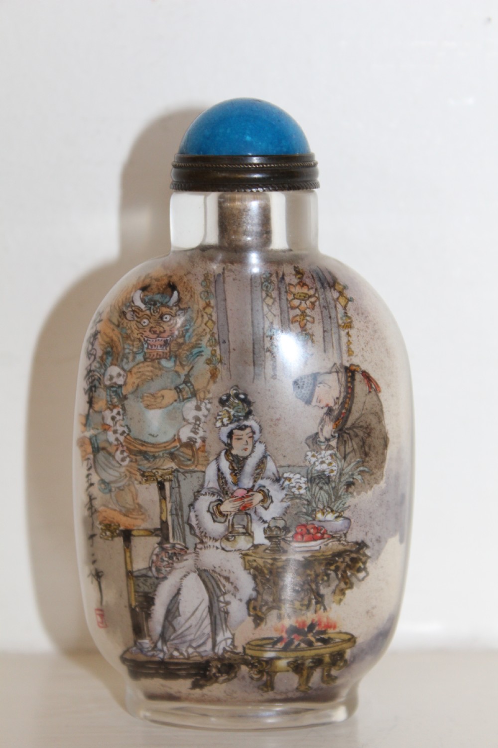 An inside-painted clear glass Chinese snuff bottle featuring detailed figures and calligraphy, topped with a rounded blue cabochon stopper. View XIII