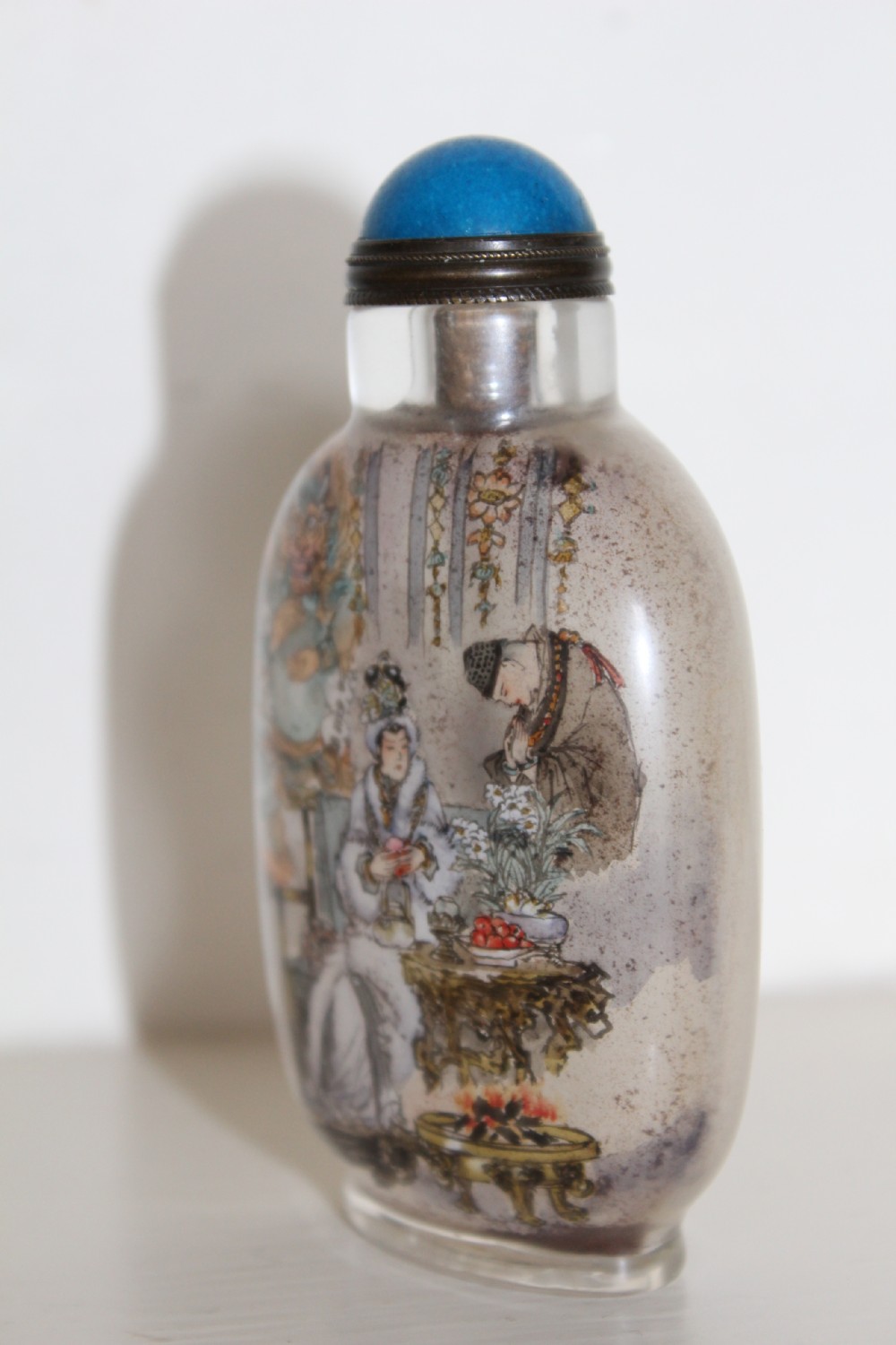 An inside-painted clear glass Chinese snuff bottle featuring detailed figures and calligraphy, topped with a rounded blue cabochon stopper. View XV