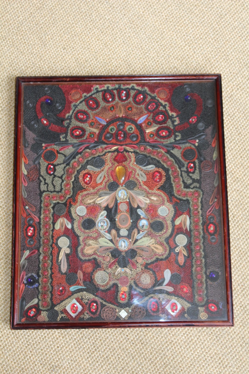 An antique framed 19th-century Indian quillwork picture featuring intricate red, black, and gold paper scrollwork embellished with beads and small painted portrait miniatures.