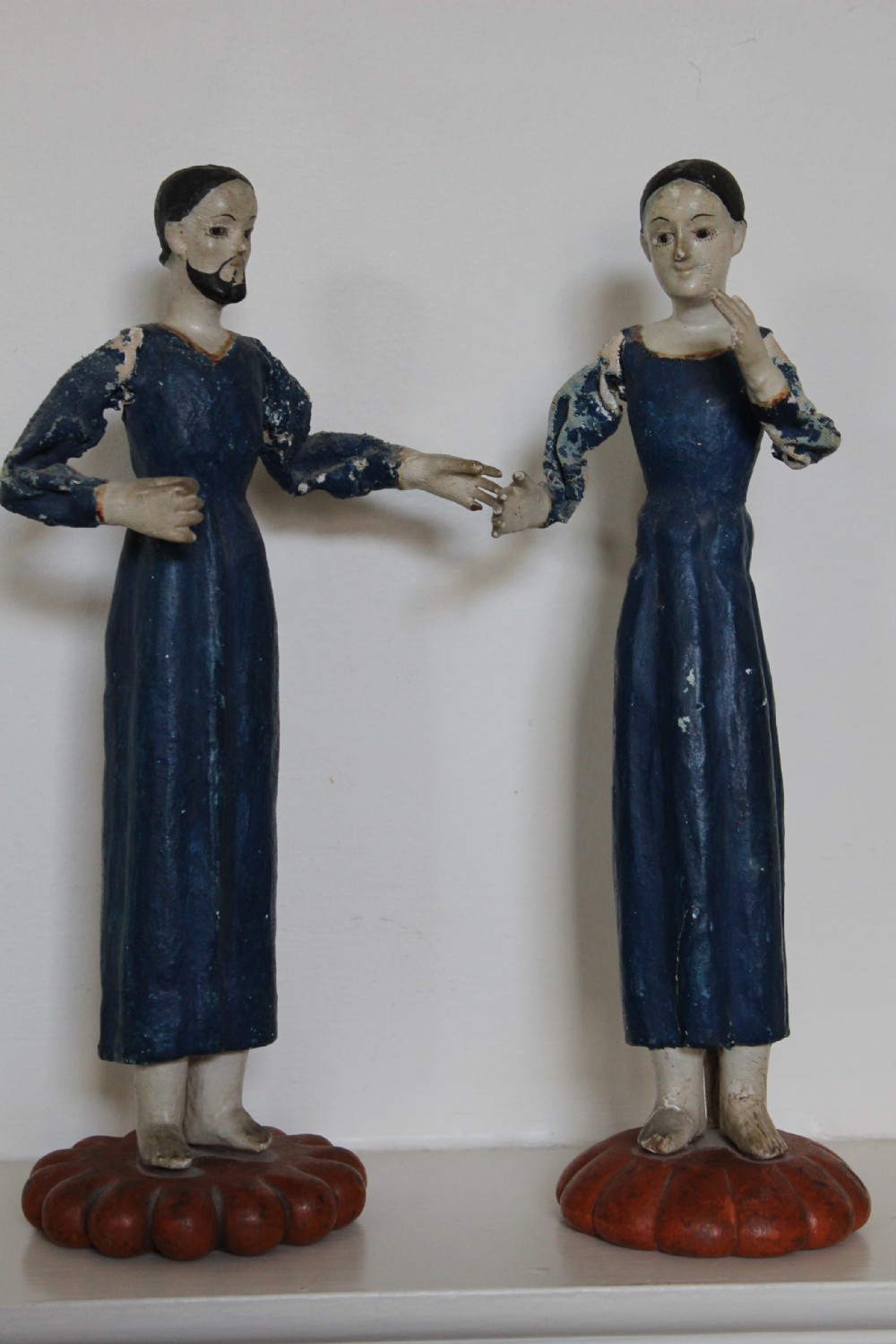 A pair of carved wooden folk art figures depicting a male and female figure dressed in blue gowns, mounted on rounded red scalloped bases.