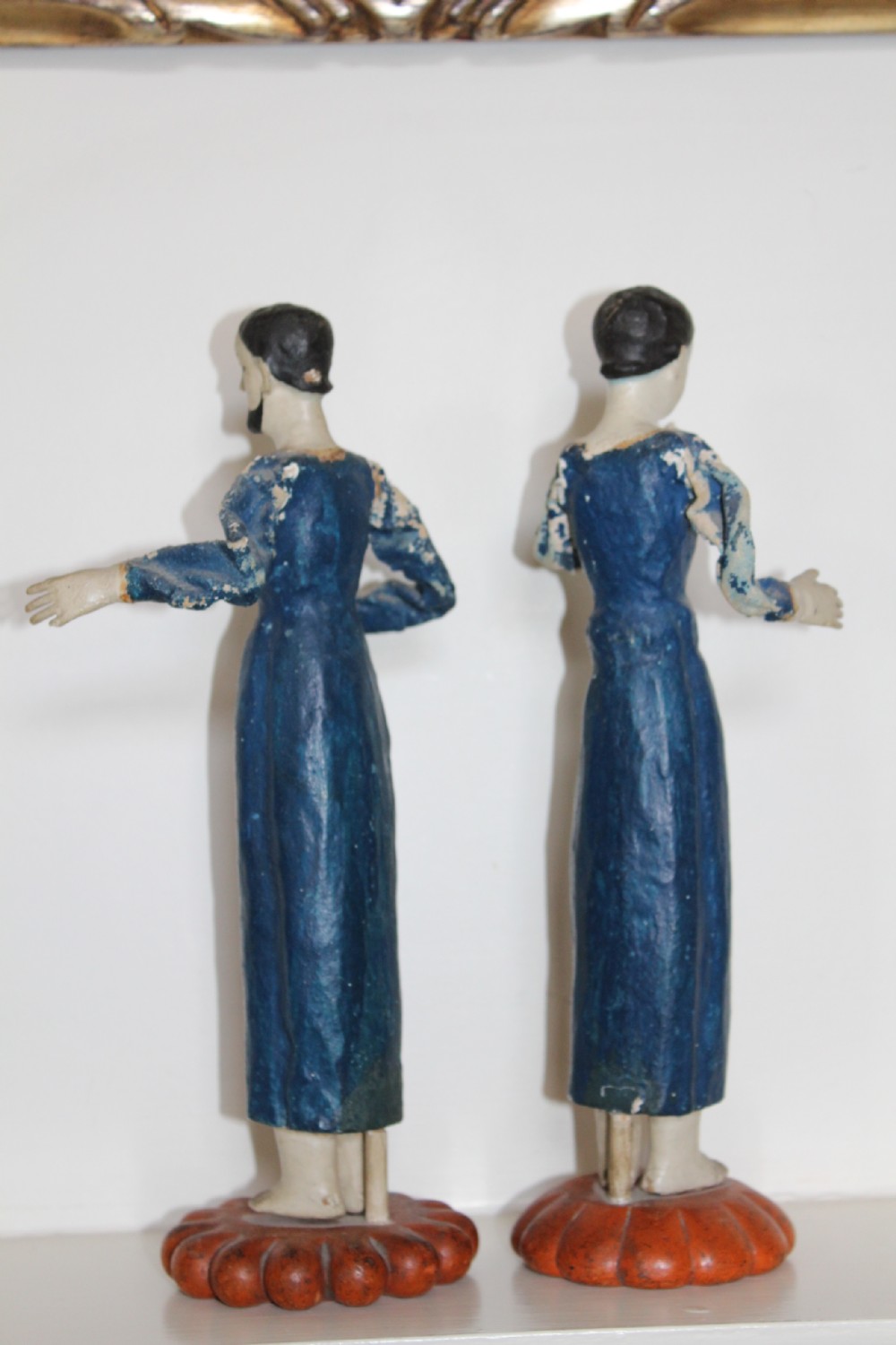 A pair of carved wooden folk art figures depicting a male and female figure dressed in blue gowns, mounted on rounded red scalloped bases. View IV