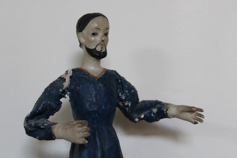 A pair of carved wooden folk art figures depicting a male and female figure dressed in blue gowns, mounted on rounded red scalloped bases. View V