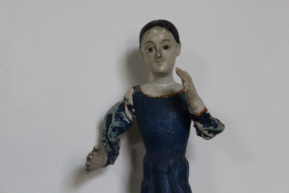 A pair of carved wooden folk art figures depicting a male and female figure dressed in blue gowns, mounted on rounded red scalloped bases. View VI