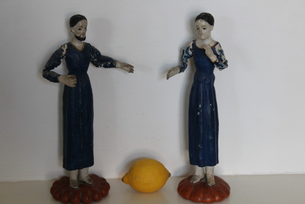 A pair of carved wooden folk art figures depicting a male and female figure dressed in blue gowns, mounted on rounded red scalloped bases. View X