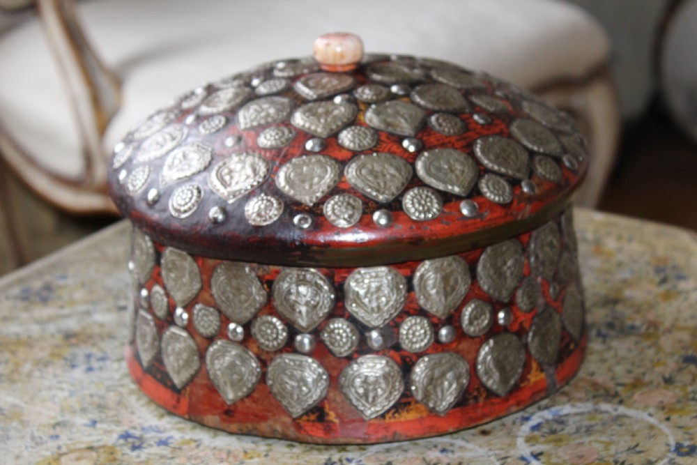 A circular red lacquered Kashmiri turban box with a domed lid, adorned with embossed silver-coloured metal mounts and a central turned knob.