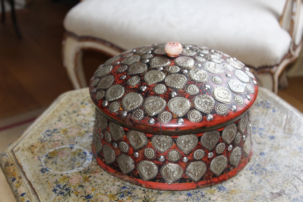 A circular red lacquered Kashmiri turban box with a domed lid, adorned with embossed silver-coloured metal mounts and a central turned knob. View VI