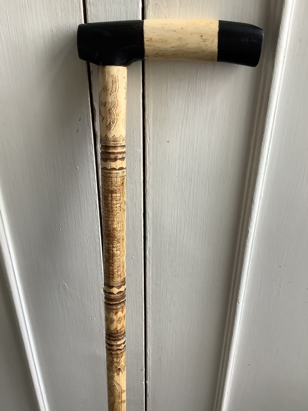 A maritime walking stick featuring a carved scrimshaw shaft with geometric patterns and a T-bar handle made of cream bone and black end caps.