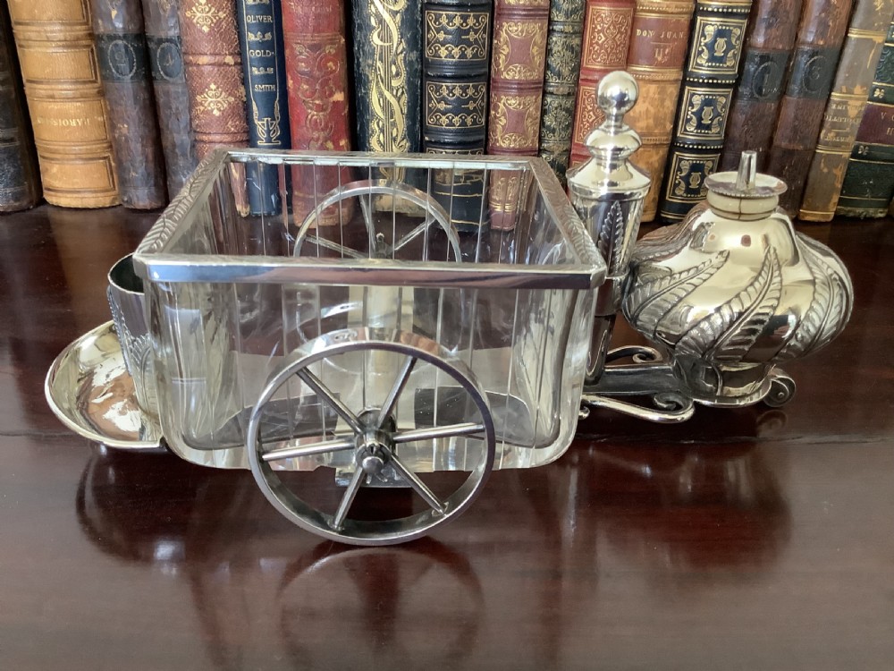 An antique silver and clear cut glass smoking compendium modelled in the form of a wheeled fire carriage with integrated lighter and storage compartments.