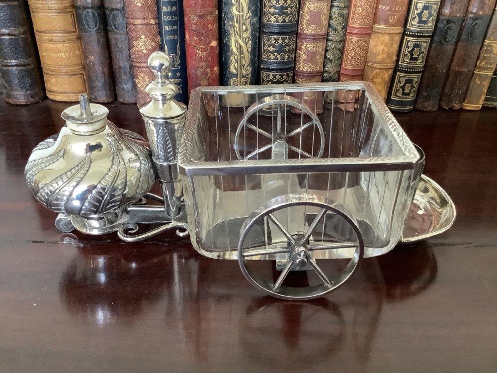 An antique silver and clear cut glass smoking compendium modelled in the form of a wheeled fire carriage with integrated lighter and storage compartments. View II