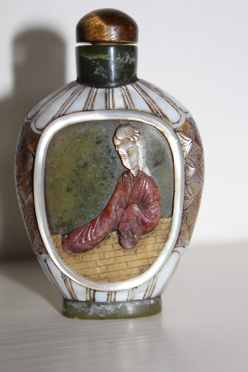A 19th-century Chinese snuff bottle featuring a carved stone figure inset within a mother-of-pearl frame on a green jade body.