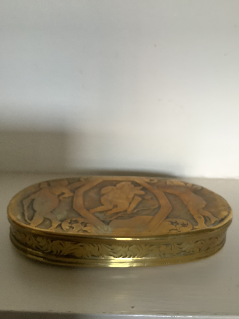 An oval heavy-gauge brass tobacco box lid featuring relief-engraved panels with cherub figures and scrollwork. View II