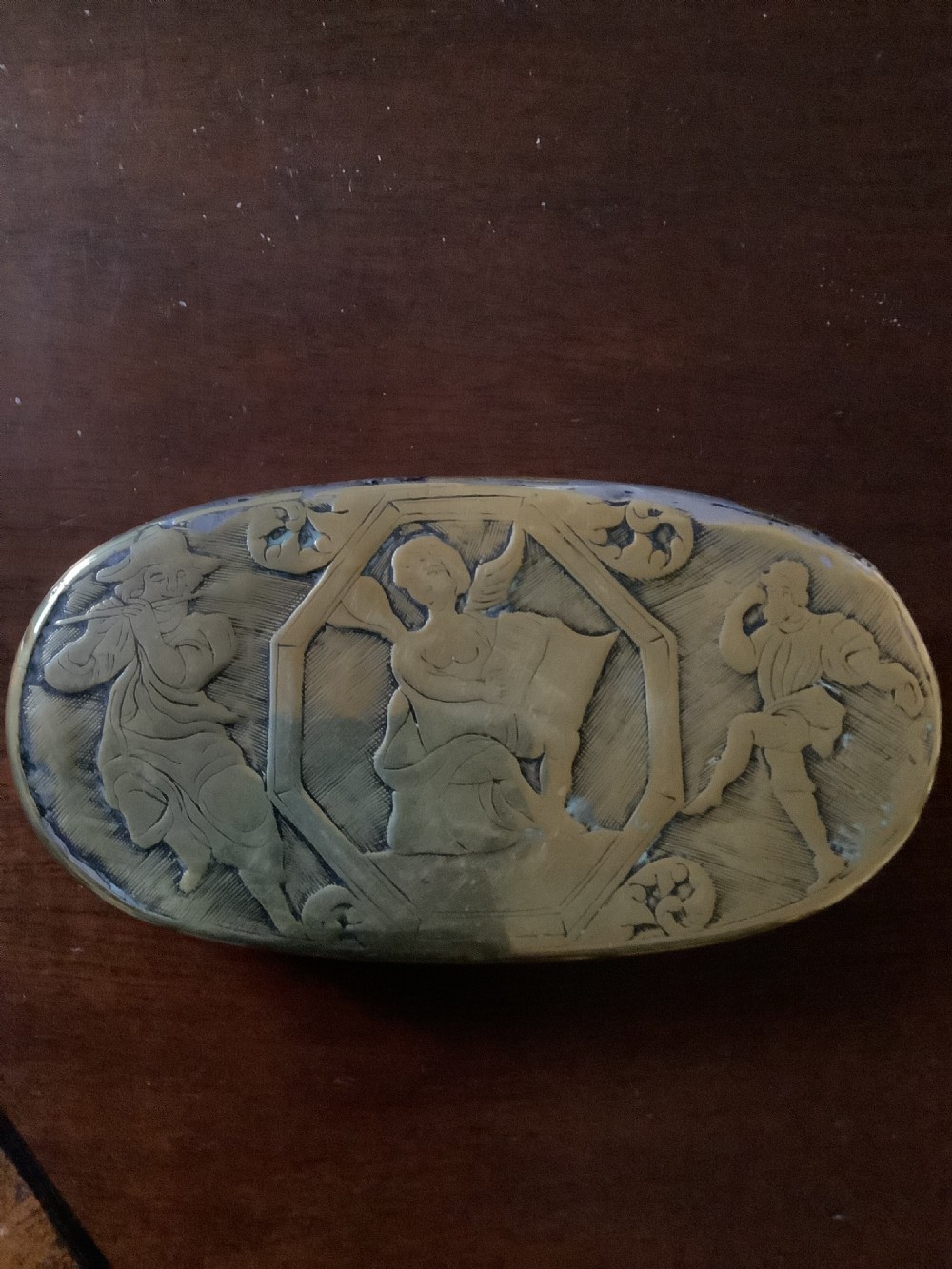An oval heavy-gauge brass tobacco box lid featuring relief-engraved panels with cherub figures and scrollwork. View IV