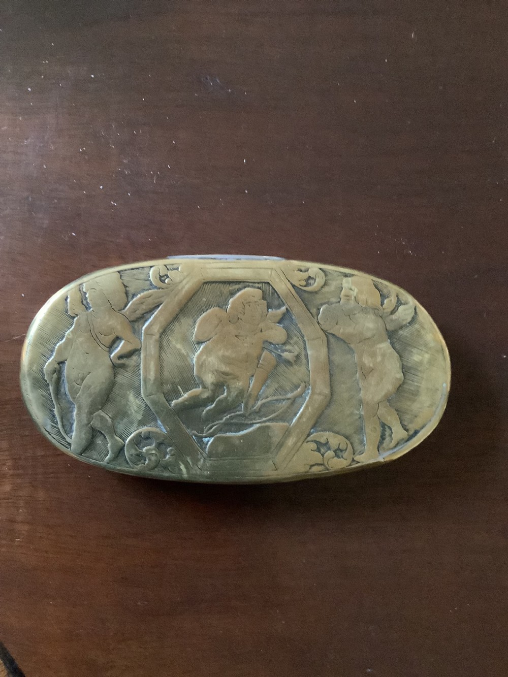 An oval heavy-gauge brass tobacco box lid featuring relief-engraved panels with cherub figures and scrollwork. View V