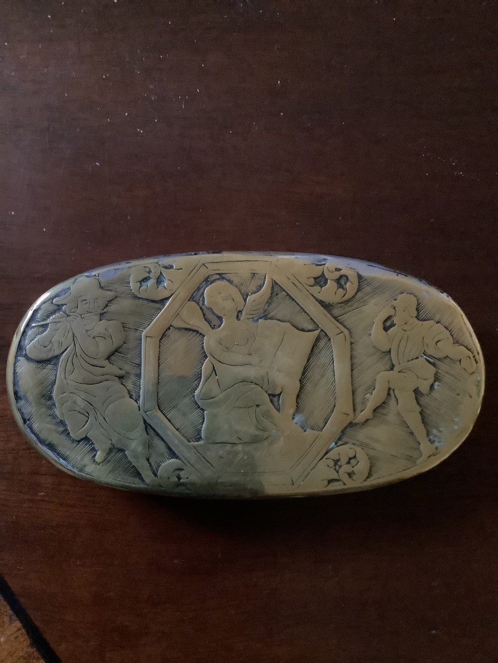 An oval heavy-gauge brass tobacco box lid featuring relief-engraved panels with cherub figures and scrollwork. View VI
