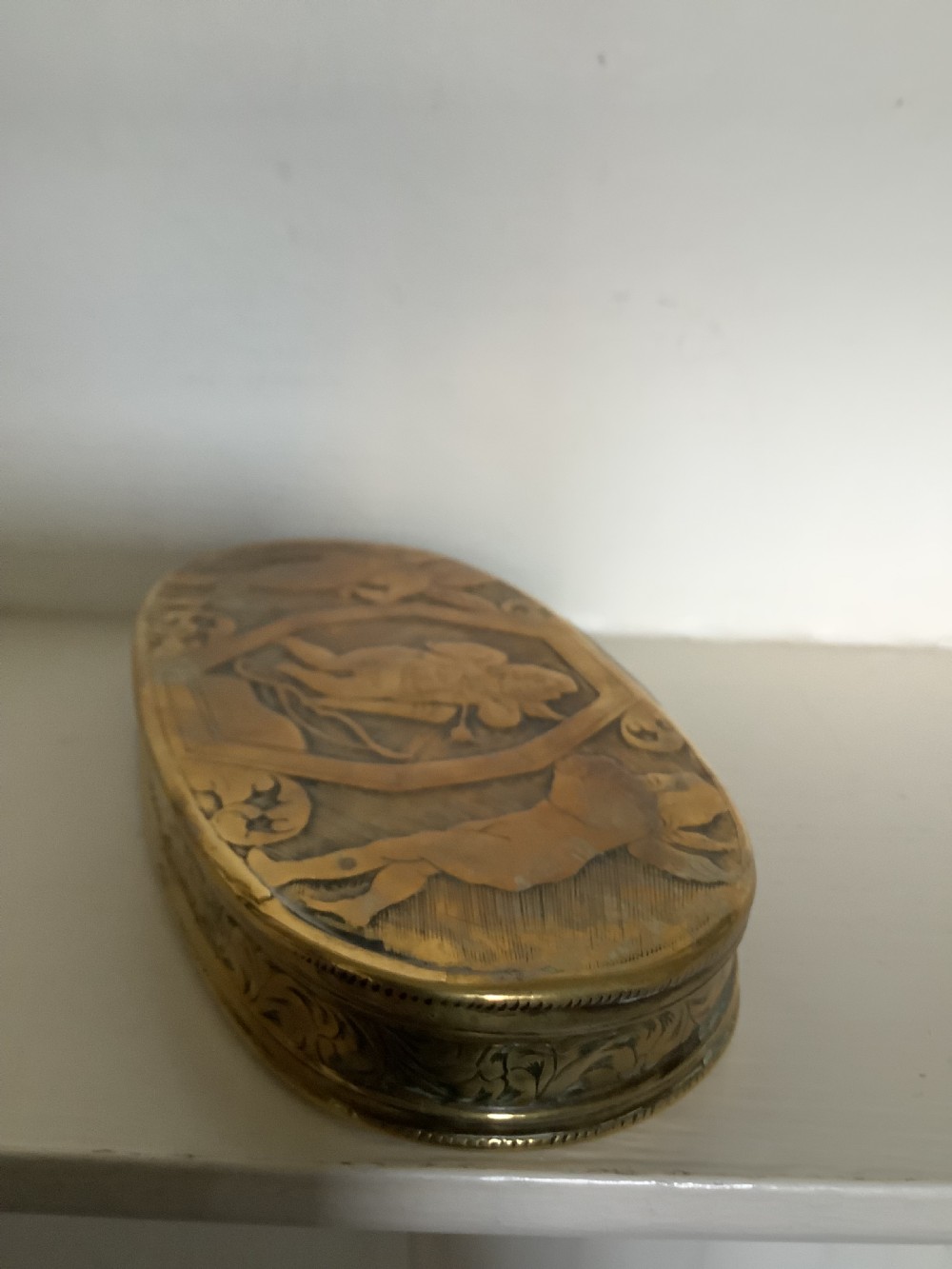An oval heavy-gauge brass tobacco box lid featuring relief-engraved panels with cherub figures and scrollwork. View VIII