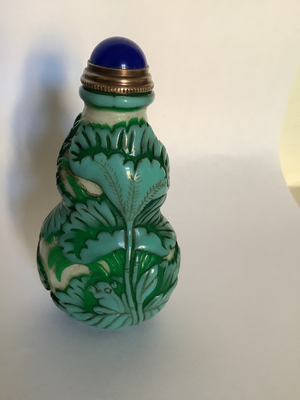 A double-gourd shaped Chinese Peking glass snuff bottle featuring carved green foliage overlay against a cream background, finished with a blue cabochon stopper. View II