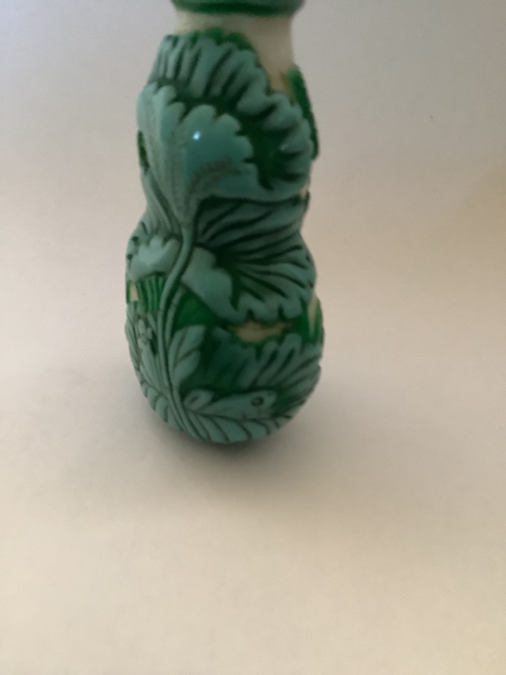 A double-gourd shaped Chinese Peking glass snuff bottle featuring carved green foliage overlay against a cream background, finished with a blue cabochon stopper. View VII