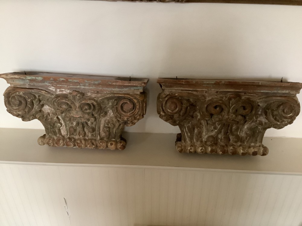 A pair of carved wooden Corinthian architectural capitals featuring acanthus leaf motifs and distressed painted finishes with metal candle inserts on top. View VI
