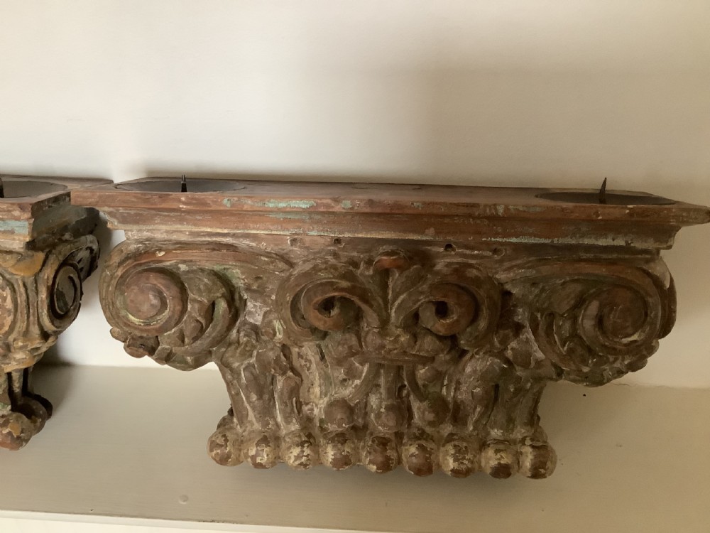 A pair of carved wooden Corinthian architectural capitals featuring acanthus leaf motifs and distressed painted finishes with metal candle inserts on top.