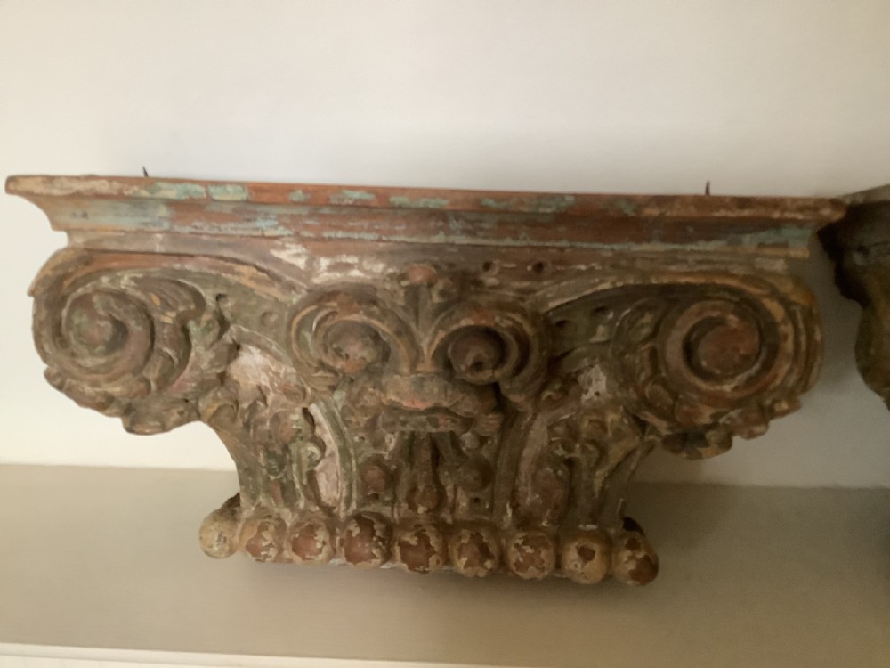 A pair of carved wooden Corinthian architectural capitals featuring acanthus leaf motifs and distressed painted finishes with metal candle inserts on top. View VII