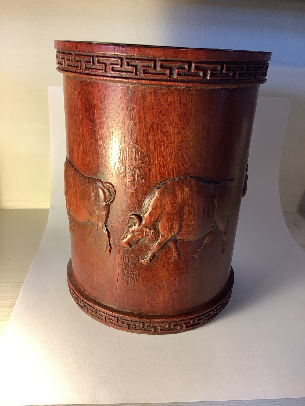 A cylindrical carved reddish brown hardwood Chinese brush pot decorated with high relief figures and an ox, bordered by geometric key fret patterns at the top and bottom edges. View IV