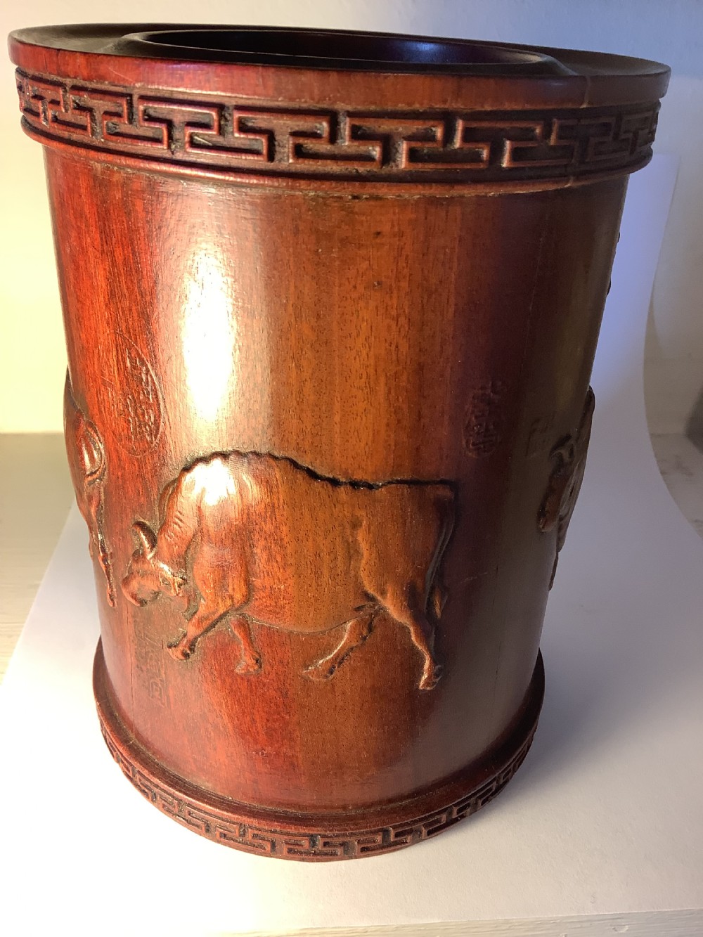 A cylindrical carved reddish brown hardwood Chinese brush pot decorated with high relief figures and an ox, bordered by geometric key fret patterns at the top and bottom edges. View V
