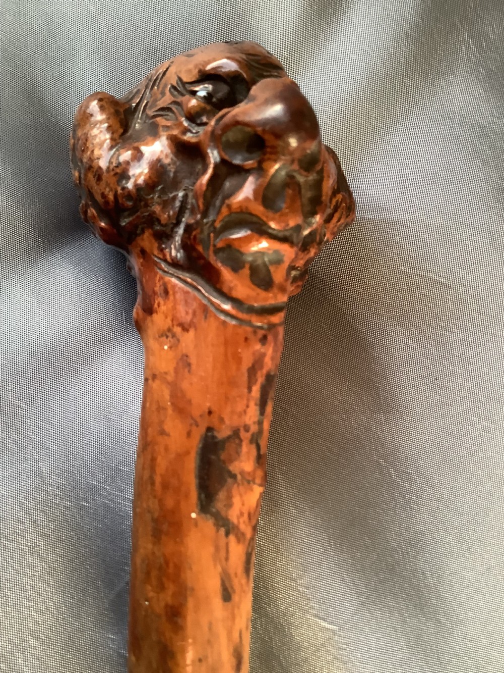 A close-up view of a warm brown hand-carved wooden stick top depicting a detailed caricature face with prominent facial features. View V