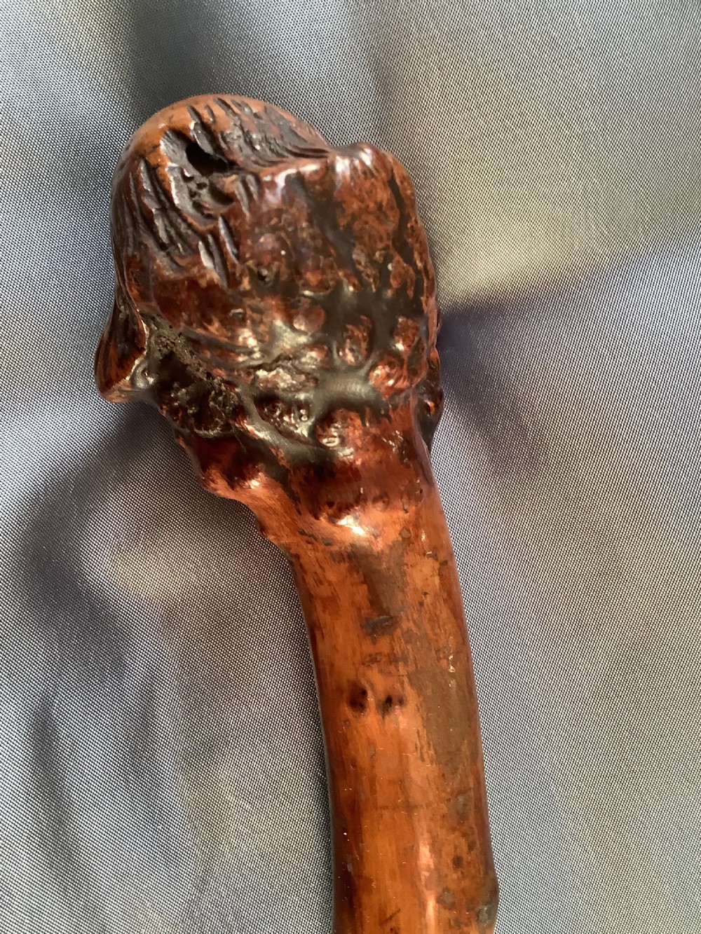 A close-up view of a warm brown hand-carved wooden stick top depicting a detailed caricature face with prominent facial features. View VI