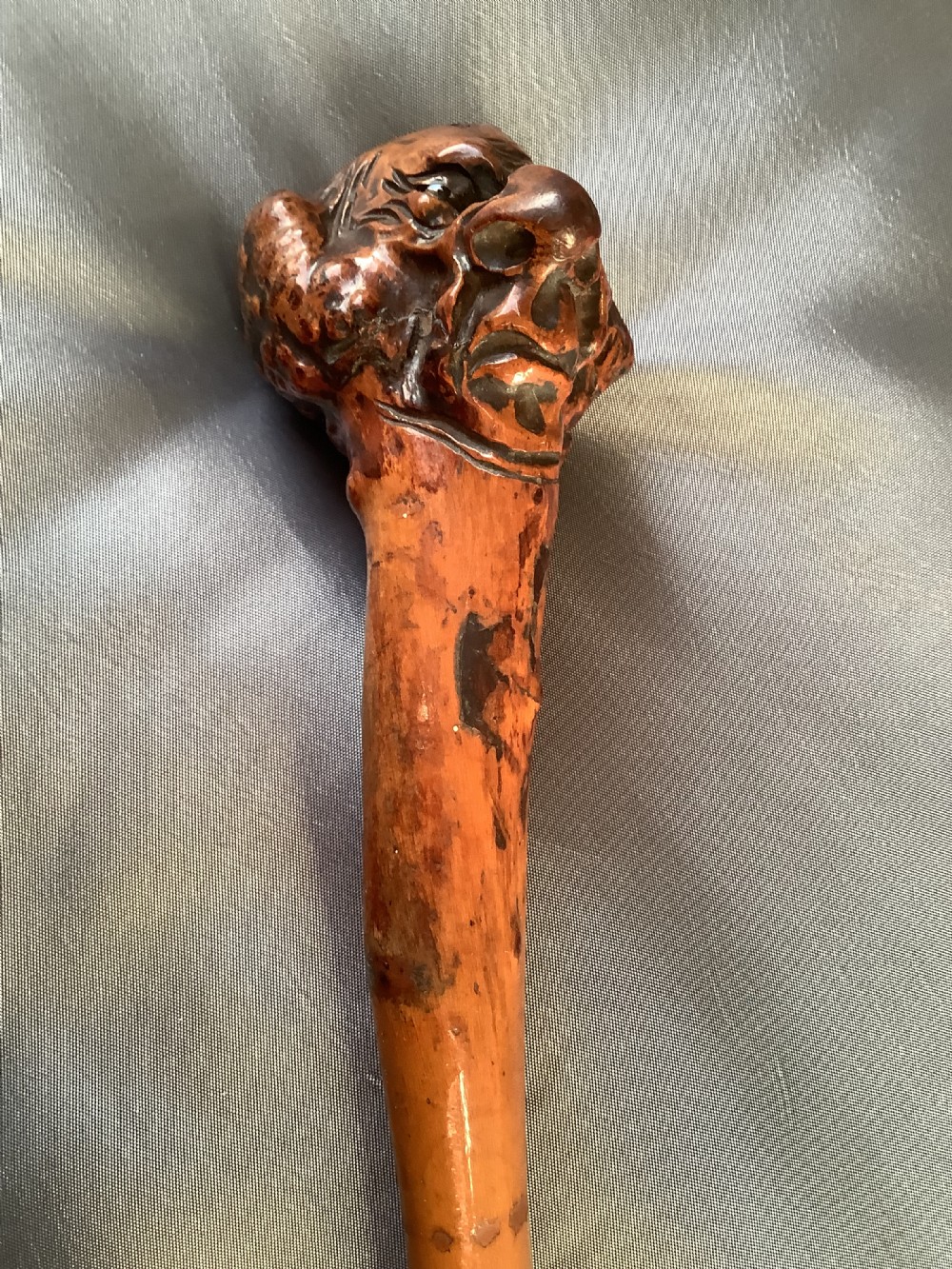 A close-up view of a warm brown hand-carved wooden stick top depicting a detailed caricature face with prominent facial features. View X