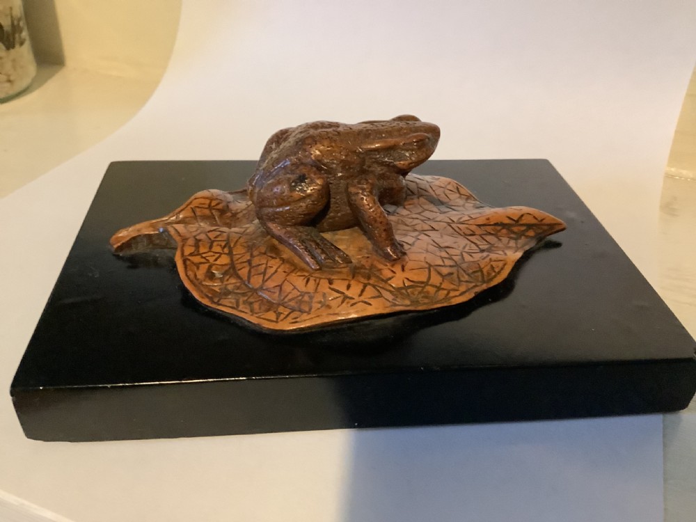 A carved brown boxwood toad sitting on an etched leaf surface mounted on a black lacquered rectangular base.