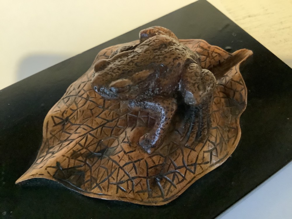 A carved brown boxwood toad sitting on an etched leaf surface mounted on a black lacquered rectangular base. View IV