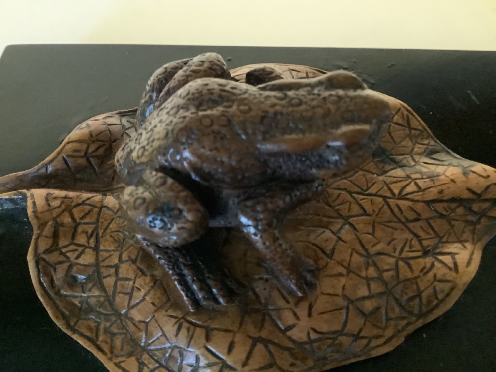 A carved brown boxwood toad sitting on an etched leaf surface mounted on a black lacquered rectangular base. View VII
