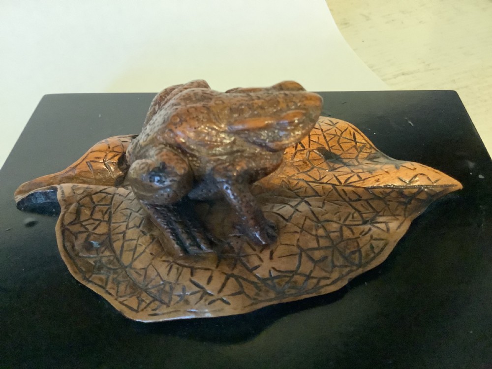 A carved brown boxwood toad sitting on an etched leaf surface mounted on a black lacquered rectangular base. View IX