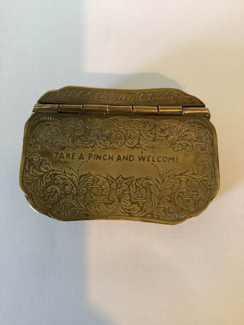 An antique hinged brass snuff box featuring ornate foliate scrollwork engraving and the central inscription reading TAKE A PINCH AND WELCOME.