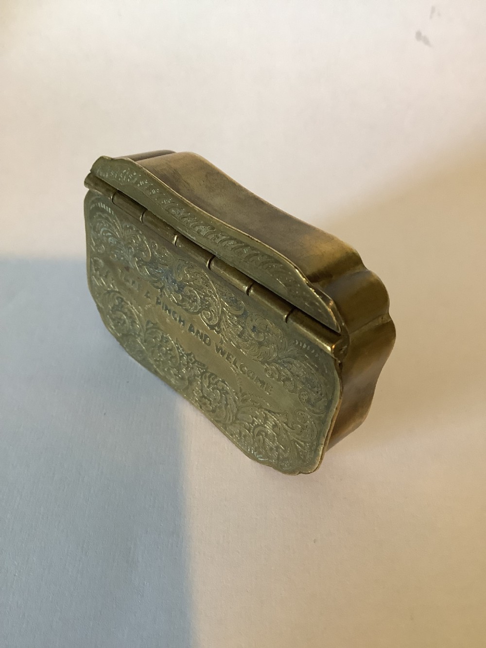 An antique hinged brass snuff box featuring ornate foliate scrollwork engraving and the central inscription reading TAKE A PINCH AND WELCOME. View IV